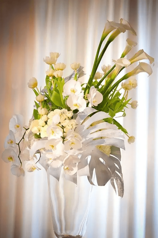 a tall tropical wedding centerpiece of tulips, orchids and callas and a large monstera leaf spray painted white looks wow