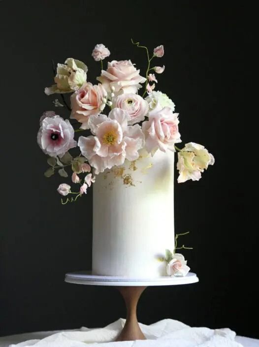 a tall white wedding cake with gold leaf, blush and white blooms covering the whole top of the cake