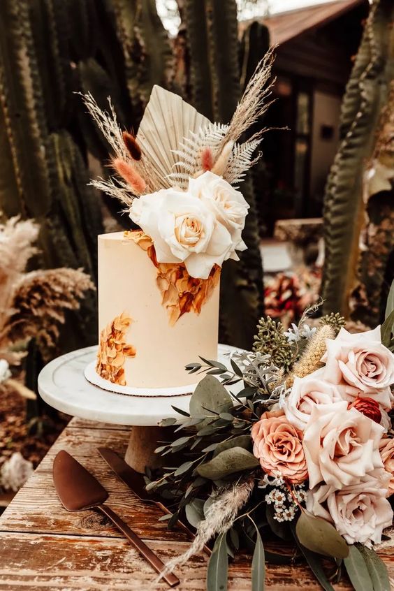 a tall white wedding cake with painted blooms, white roses, fronds, bunny tails, grasses for a fall boho wedding