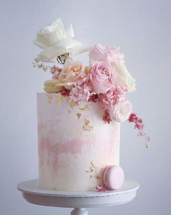 a watercolor blush wedding cake topped with pink and white roses and macarons and accented with gold leaf