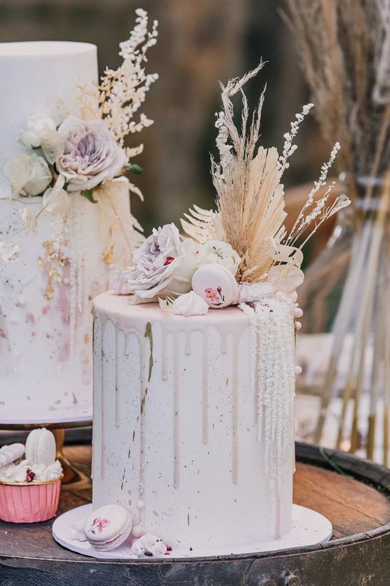 a white tall wedding cake with pale pink drip, matching roses, painted macarons and pampas grass and fronds