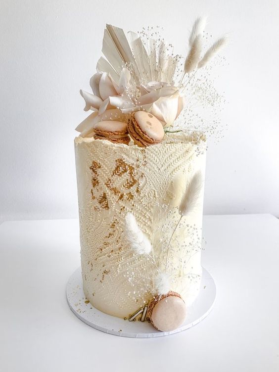 a white textural wedding cake with white fronds, bunny tails, pink macarons and some baby’s breath