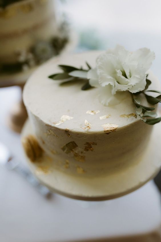 a white textured wedding cake topped with gold foil and a white bloom and greenery is a chic and cool idea