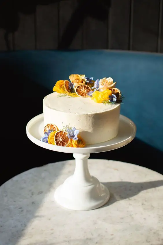 a white wedding cake topped with fresh yellow and blue blooms adn dried citrus slices is a cool idea for a summer or fall wedding