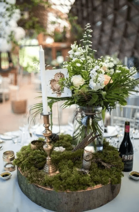 an enchanted forest wedding table setting with a wood slice, moss, a tall and thin candle, a jar with greenery and neutral blooms and feathers
