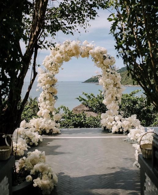 a super lush white floral wedding arch with a sea view and matching white blooms lining up the aisle is amazing and cool