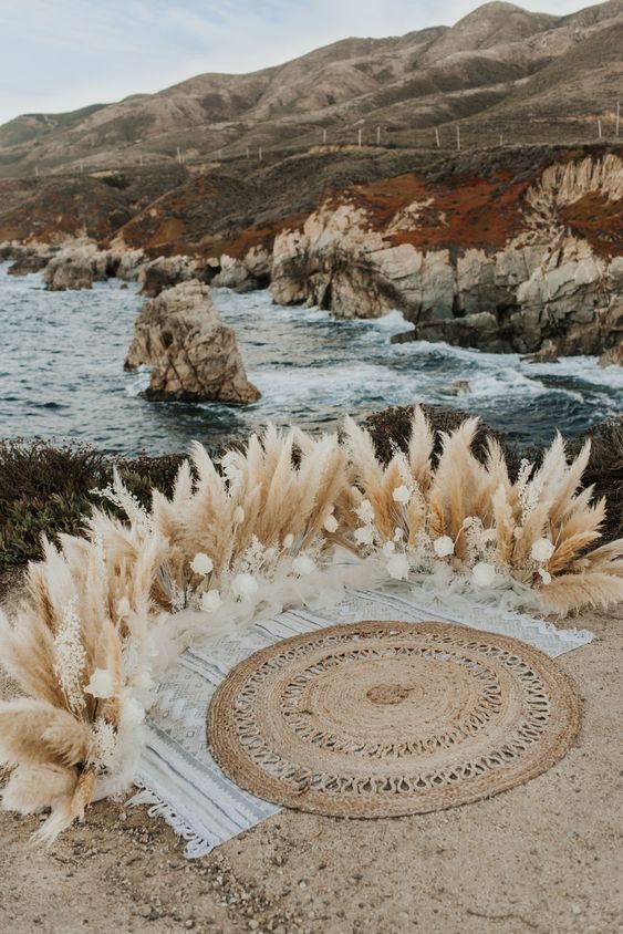 a boho wedding altar on the ground with pampas grass and white roses plus some boho rugs and a cool sea view is wow