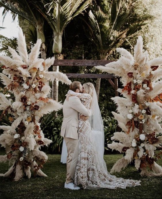 a boho wedding arch covered with pampas grass, fronds, bold blooms is a cool idea for a boho tropical or fall wedding