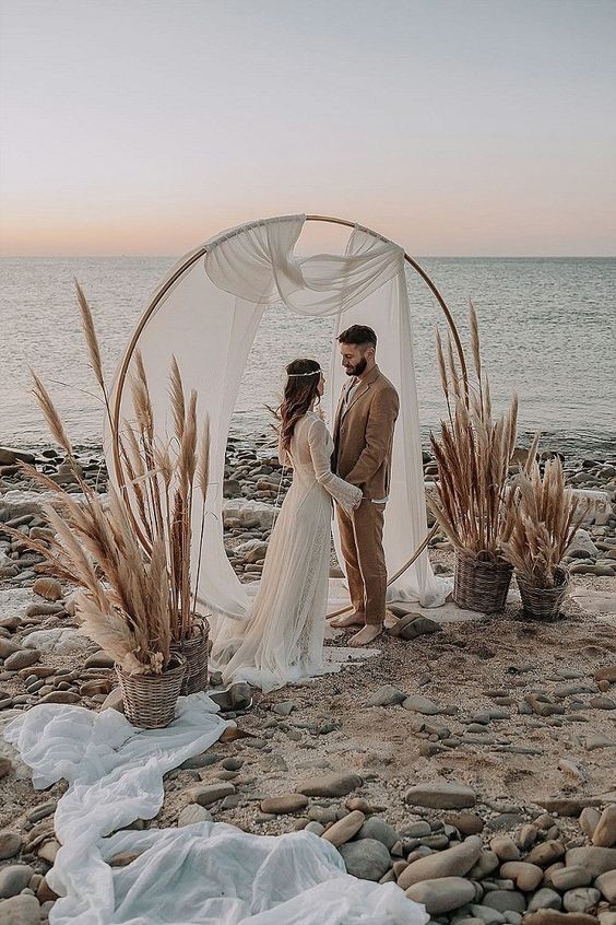 a boho wedding arch covered with white fabric and pampas grass in baskets is a very creative and unique decor idea