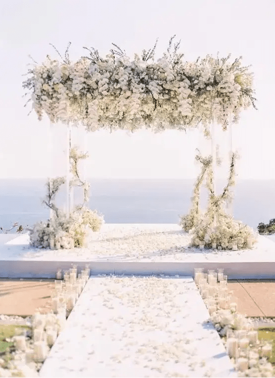 a breathtaking wedding ceremony space with a sea view, an acrylic arch with lush white florals, pillar candles lining up the aisle