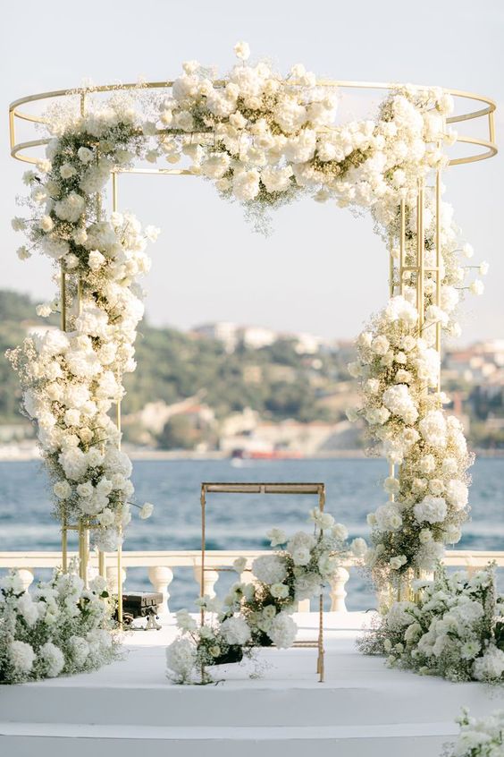 a chic white wedding arch with some roses, hydrangeas and baby's breath plus a gorgeous sea view