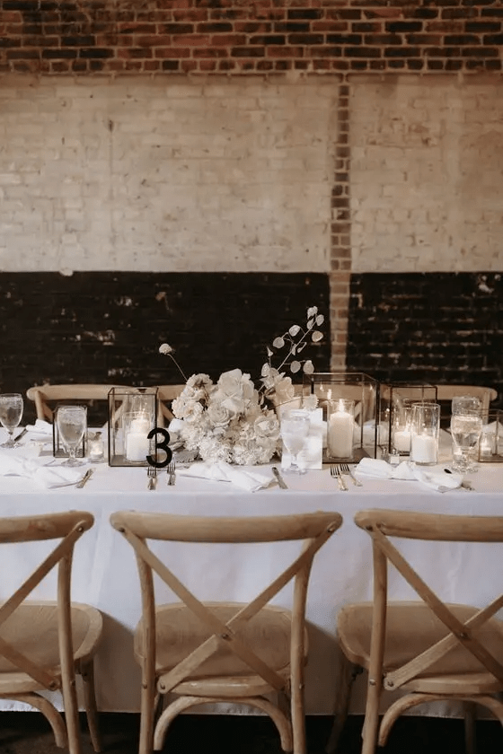 a chic winter wedding tablescape done in white, with candle lanterns, a lush white floral centerpiece, a black table number
