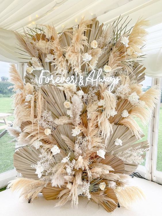 A gorgeous boho wedding backdrop composed of fronds, pampas grass, white blooms and a neon sign looks jaw dropping