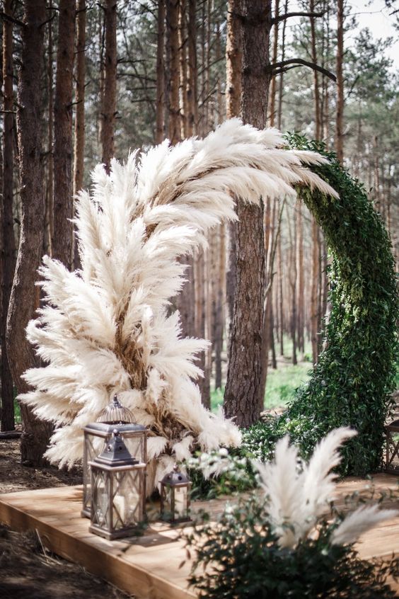 a gorgeous round boho wedding arch covered with pampas grass and greenery and lanterns at the base is amazing for a boho wedding