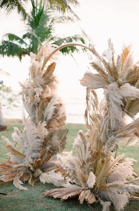 a lush round boho wedding arch covered with fronds and pampas grass is a cool idea for boho, tropical and many other weddings
