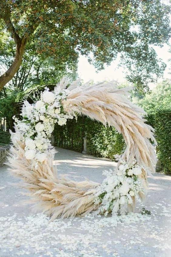 a lush round wedding arch covered with pampas grass and white blooms and greenery is amazing for spring and summer