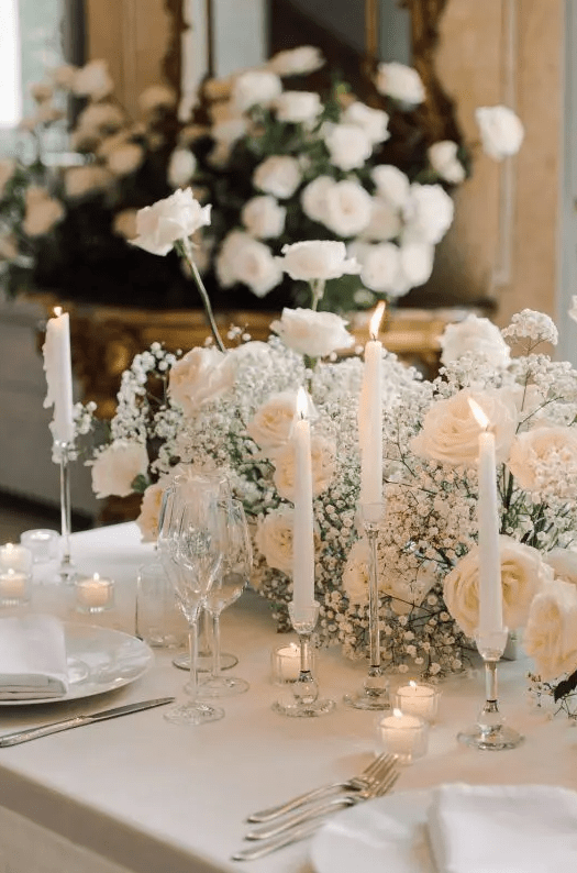 a modern romantic white wedding centerpiece of baby’s breath and roses plus tall and thin and smaller candles is pure elegance and chic