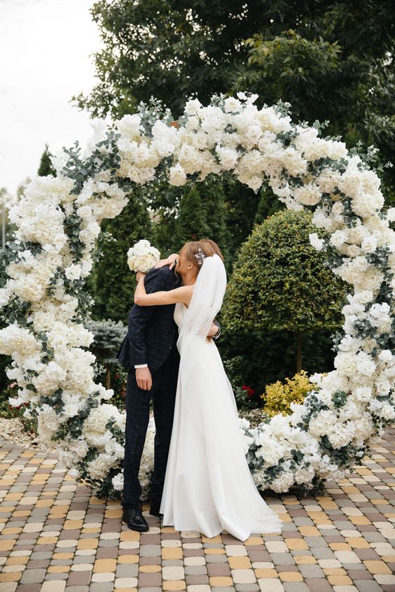 a round wedding arch decorated with white roses and hydrangeas and some greenery is a stylish and cool idea for spring and summer