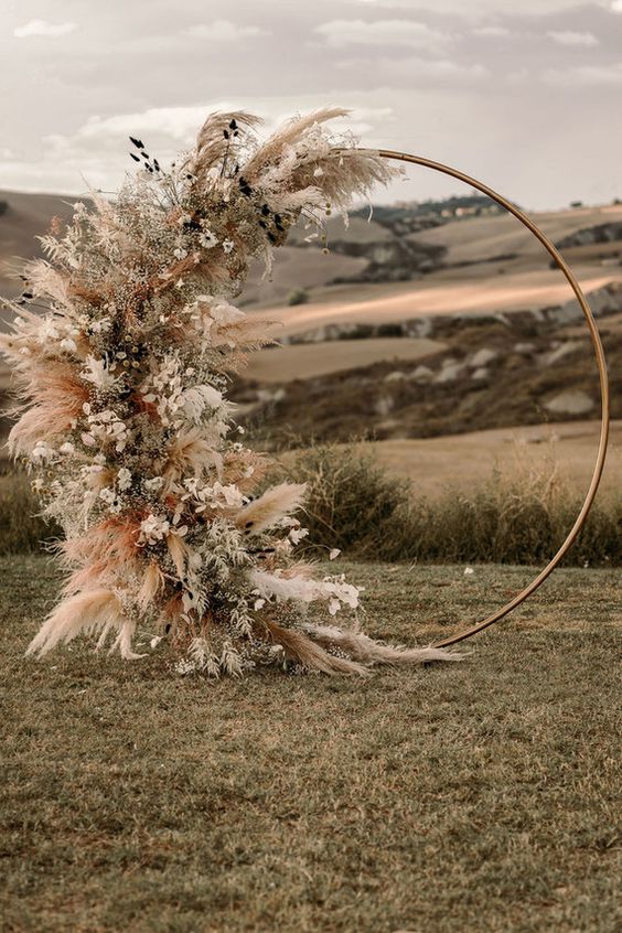 a round wedding arch partly covered with greenery, pampas grass, white and dried blooms and some dried leaves