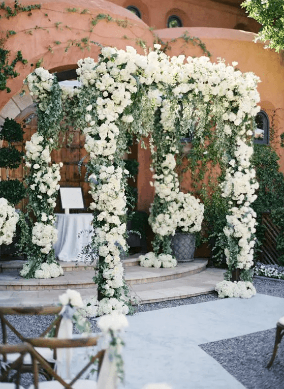 a secret garden wedding altar decorated with greenery and white blooms and with blooms at the base is a very chic and stylish solution