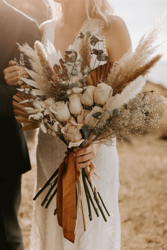 a stylish boho wedding bouquet of white roses, greenery, dried leaves and fronds and pampas grass is wow