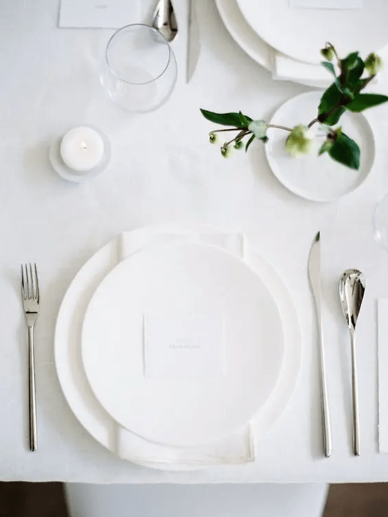 a white all-minimalist wedding tablescape with all white everything, some greenery to refresh the look and candles