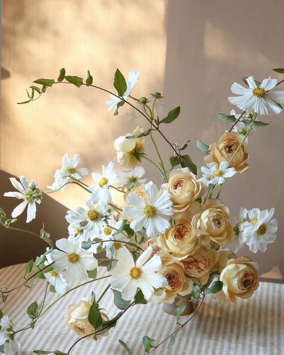 an airy summer wedding centerpiece of uellow roses and white cosmos plus greenery is adorable