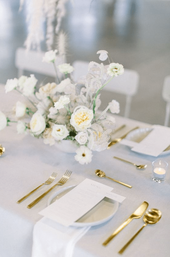 an ethereal spring wedding tablescape with neutral linens, gold cutlery, a white floral centerpiece and white stationery