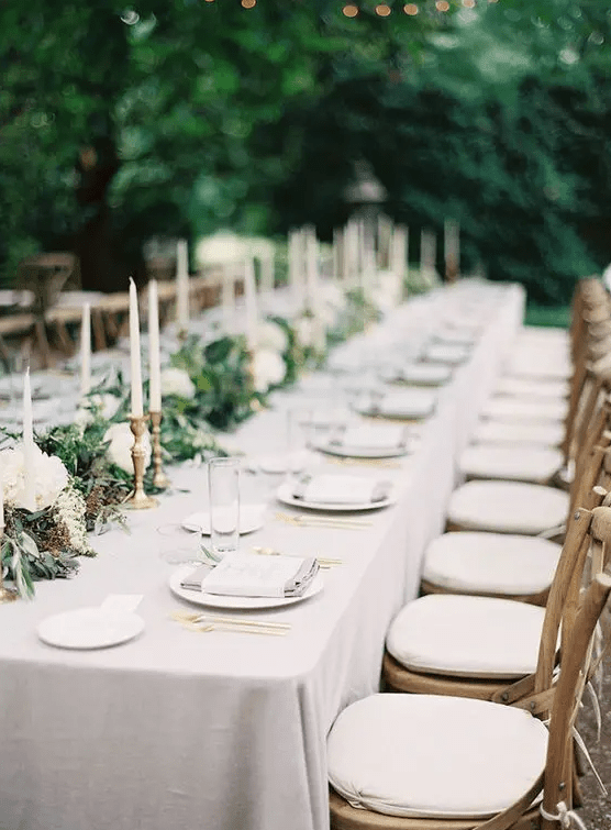 an outdoor neutral wedding tablescape with a greenery and white bloom runner, candles, porcelain and some napkins