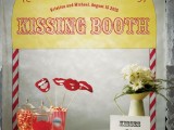 a bright kissing booth and dessert table in one, with flowers, a sign, props and lots of tasty sweets