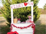 a white pallet kissing booth with bright pillows, signs, a fabric bunting, red balloons doubles as a dessert table with red sweets