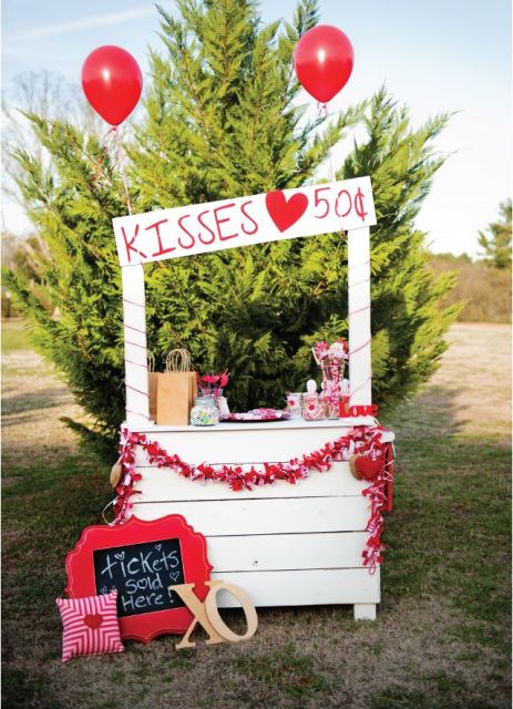 a white pallet kissing booth with bright pillows, signs, a fabric bunting, red balloons doubles as a dessert table with red sweets