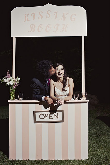 a simple and cute wedding kissing booth done in pink and white stripes, some blooms and a sign over it