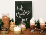 a cozy and cute hot chocolate bar with a chalkboard sign, glittered jars with sweets and candies and evergreens
