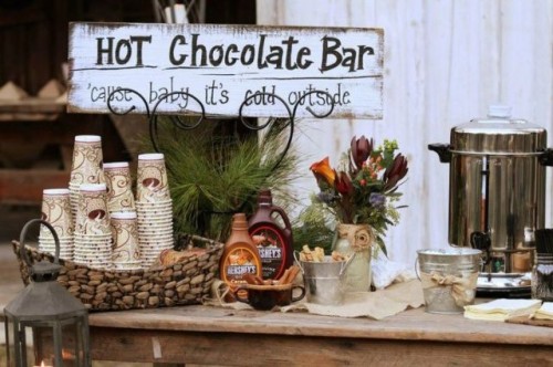 a small rustic hot chocolate bar with evergreens, dark blooms, a basket with cups and chocolate sauce