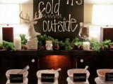 a cozy hot chocolate bar done with evergreens, a deer, a large sign, pinecones, cups and toppings of various kinds