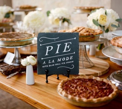 an elegant pie bar with white blooms and greenery, with a chalkboard sign, pies on glass and metal stands is very chic