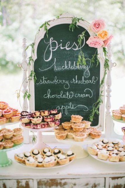 a simple vintage pie bar with a chalkboard sign decorated with greenery and blooms and with various stands with pies is a cool and lovely idea