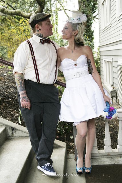 a vintage groom's outfit with grey pants, suspenders, an ivory shirt, a printed bow tie and a cap, cuffed sleeves highlight the tattoos