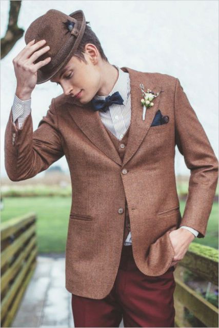 a vintage-inspired groom's look with an ocher waistcoat and jacket, red pants, a striped shirt, a navy bow tie and a brown hat
