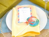 a colorful place setting with a clear plate and a bold yellow napkin, a brightmenu and a colorful lollipop for a fun and cool wedding