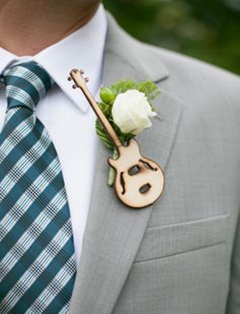 a wedding boutonniere of a wood burnt guitar, a white bloom and greenery for a music-loving groom