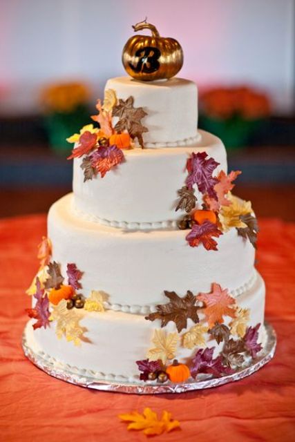 a white buttercream wedding cake with colorful faux leaves and a gold pumpkin topper is a lovely and fun idea