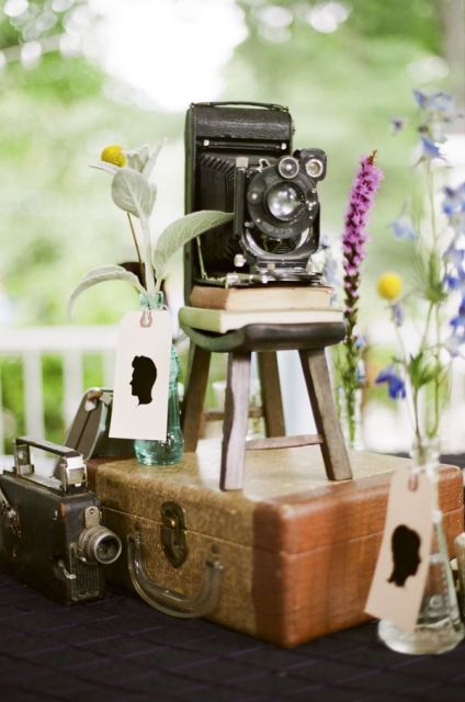 a vintage wedding centerpiece of a vintage suitcase, vintage cameras, with bold blooms and greenery in test tubes is a chic idea