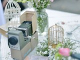 a vintage cluster wedding centerpiece of a camera, cages, books, greenery and pink flowers is a cool solution for a wedding