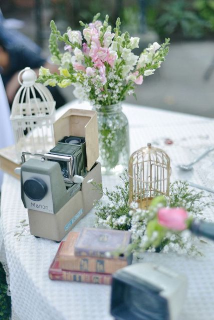 a vintage cluster wedding centerpiece of a camera, cages, books, greenery and pink flowers is a cool solution for a wedding
