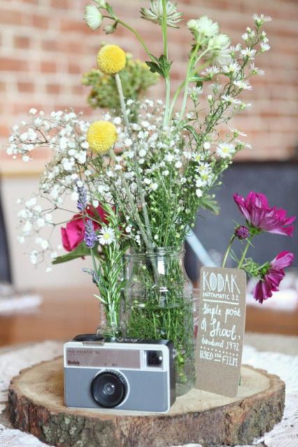 a vintage cluster wedding centerpiece of a wood slice, a jar with wildflowers and a vintage camera is ideal for a rustic wedding