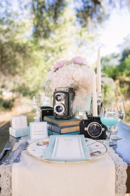 a refined wedding centerpiece composed of a stack of books, blush and white blooms, a vintage camera and tall and thin candles