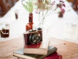 a cluster wedding centerpiece of a stack of books, a vintage camera and a beer bottle with wildflowers is lovely
