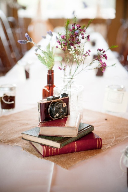 a cluster wedding centerpiece of a stack of books, a vintage camera and a beer bottle with wildflowers is lovely